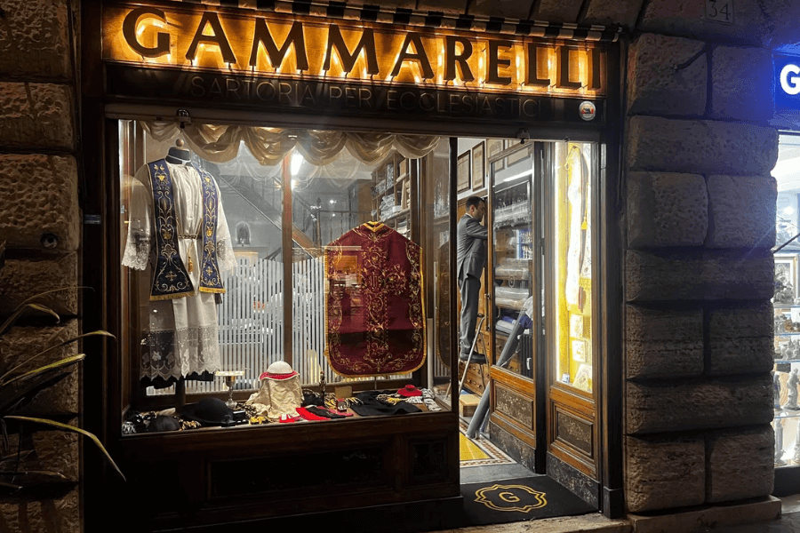 Nighttime view of the Gammarelli shopfront in Rome, a renowned ecclesiastical tailor, with ornately embroidered vestments, lace garments, birettas, and other clerical accessories displayed in the warmly lit window; a tailor is seen working inside.