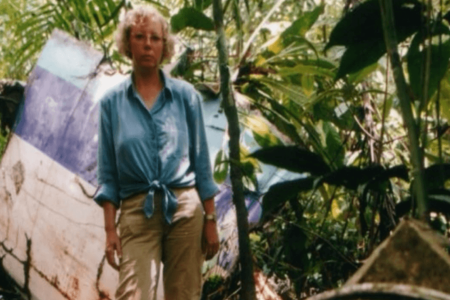 A woman stands solemnly in a dense jungle, wearing a blue button-up shirt and khaki pants, with an old, weathered aircraft wreckage behind her. The metal debris, partially overtaken by plants, suggests a long-past crash site.