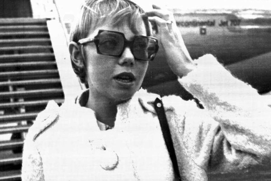 Black and white photo of a woman in oversized sunglasses and a textured coat, standing in front of an airplane staircase with part of the aircraft visible behind her. She is adjusting her hair and appears to be speaking or mid-sentence.