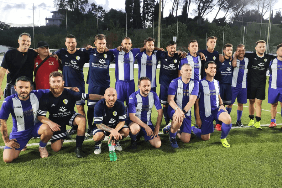 Group photo of two soccer teams posing together on a field after a match, with players wearing coordinated navy blue and purple kits bearing logos such as “Bambino Gesù” and “Studium Parthenon,” under evening light with trees and fencing in the background.