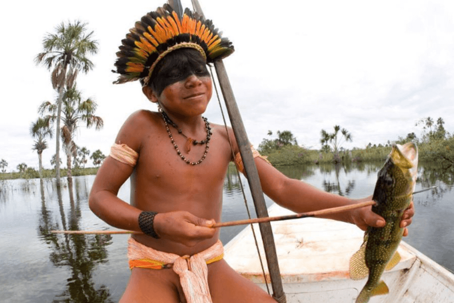 A smiling Indigenous boy wearing traditional attire, body paint, and a vibrant feathered headdress proudly holds up a freshly caught fish while sitting in a canoe on a calm, reflective river surrounded by palm trees.