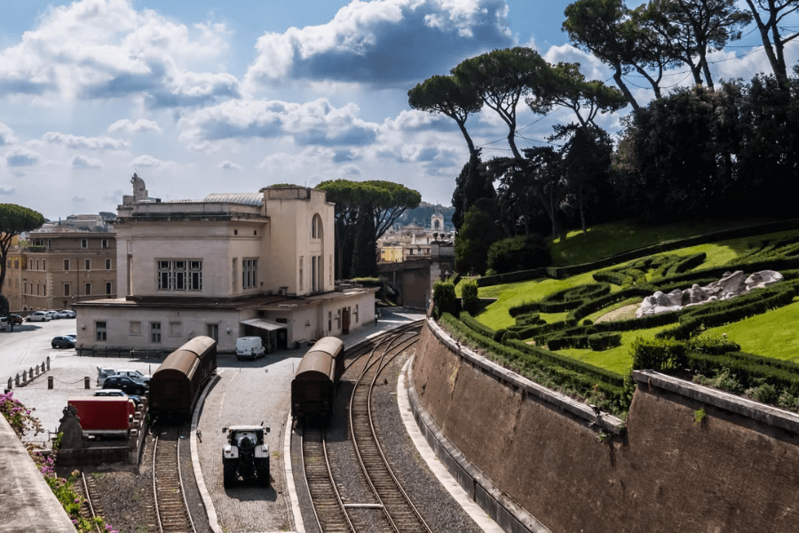 Curving railway tracks lead into Vatican City, passing by a cream-colored train station building and flanked by manicured gardens and sculpted hedges on one side, with tall pine trees and the city of Rome visible in the background under a partly cloudy sky.