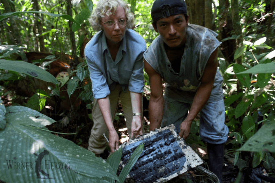 A woman and a man in a dense jungle bend down to lift a weathered and partially decayed piece of aircraft wreckage, likely a control panel. The surrounding foliage is thick and green, suggesting a tropical environment, with visible tension and focus on their faces.