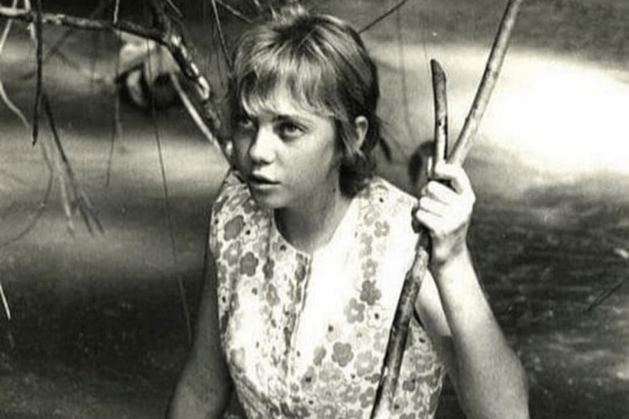 Black-and-white photo of a young girl with short hair and a floral-patterned sleeveless dress, standing in shallow water while holding onto branches. She looks alert and focused, possibly navigating through a forested or jungle area.