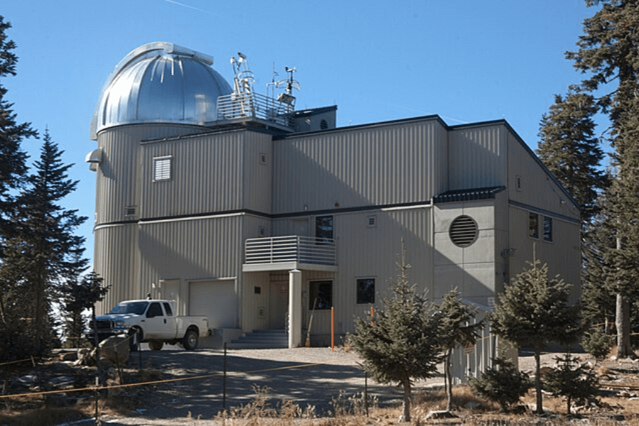 Exterior view of the Vatican Advanced Technology Telescope (VATT) facility, a metal-clad observatory building with a domed roof housing the telescope, located among pine trees on Mount Graham in Arizona, with a white truck parked out front.