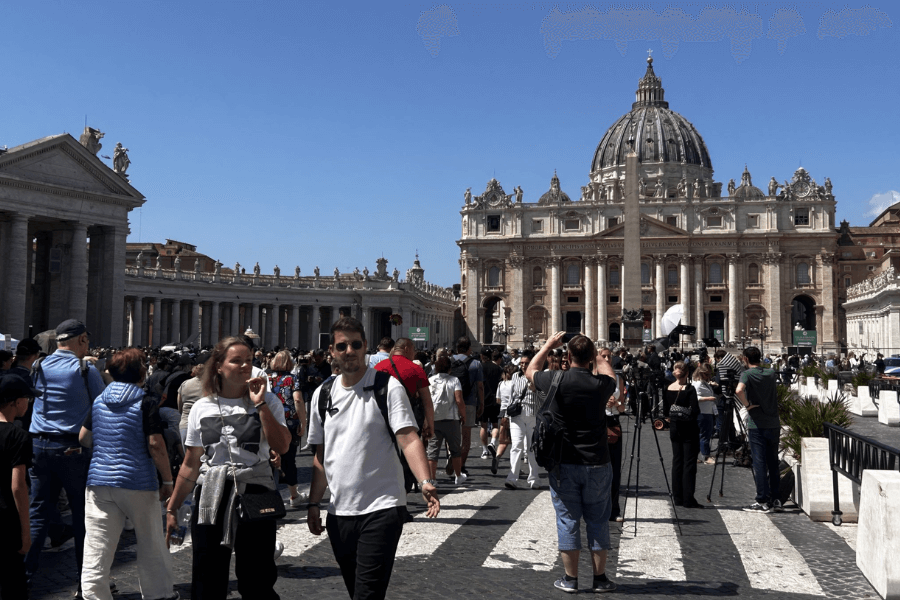 Crowds of tourists and media personnel with cameras gather in St. Peter’s Square under a clear blue sky, facing St. Peter’s Basilica with its grand façade and iconic dome, while the central obelisk stands prominently in front.