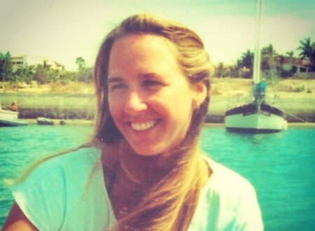 Tami Oldham’s Unbelievable Fight to Survive After a Storm Took Everything Young woman smiling brightly while sitting on a boat in turquoise water, with a dock, sailboat, and palm trees visible in the sunlit background. Her long blonde hair is gently windswept, and she wears a white shirt under the warm, tropical light.