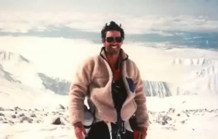 The Man Who Came Back from the Dead: Beck Weathers’ Shocking SurvivalBeck A man wearing sunglasses and a fleece jacket smiles while standing on a snowy mountain summit with a vast, rugged landscape of snow-covered peaks behind him.