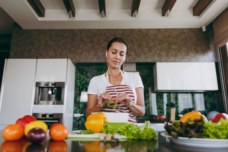 Young woman with headphones in the ears holding vegetables in hands in kitchen with laptop on the table. Vegetable salad. Dieting concept. Healthy lifestyle. Cooking at home. Prepare food