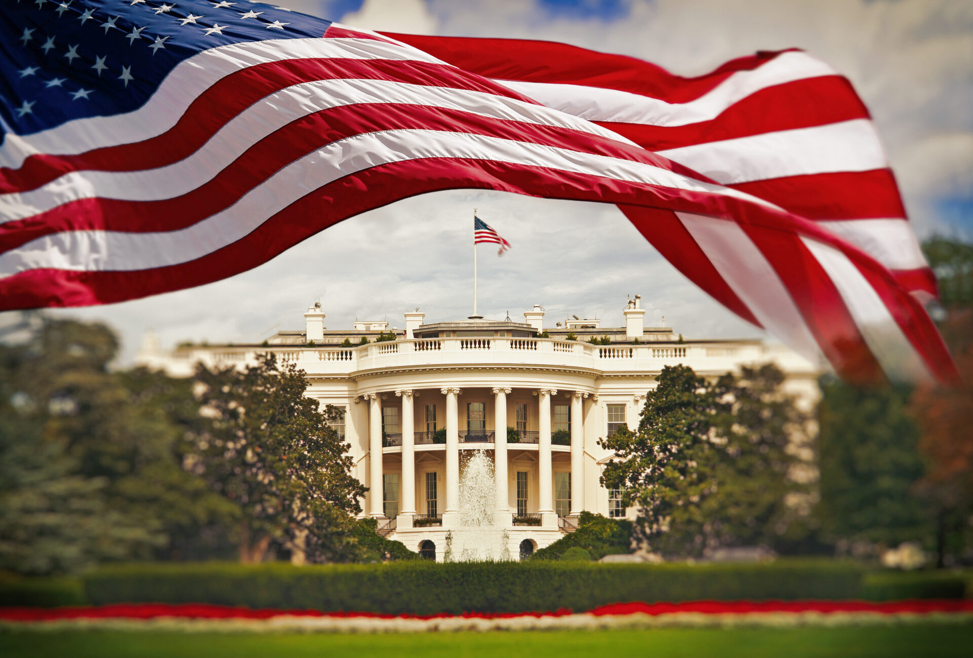 American flag waving in foreground with the White House in Washington, D.C. in the background.