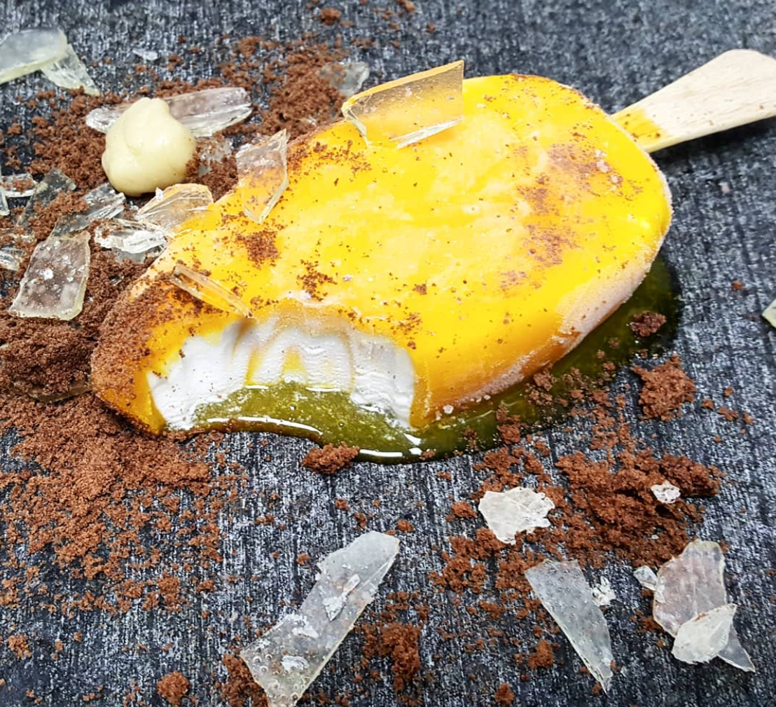 Orange creamsicle with one bite out of it on the pavement surrounded by broken glass and dirt, edible optical illusion by chefbenchurchill on instagram
