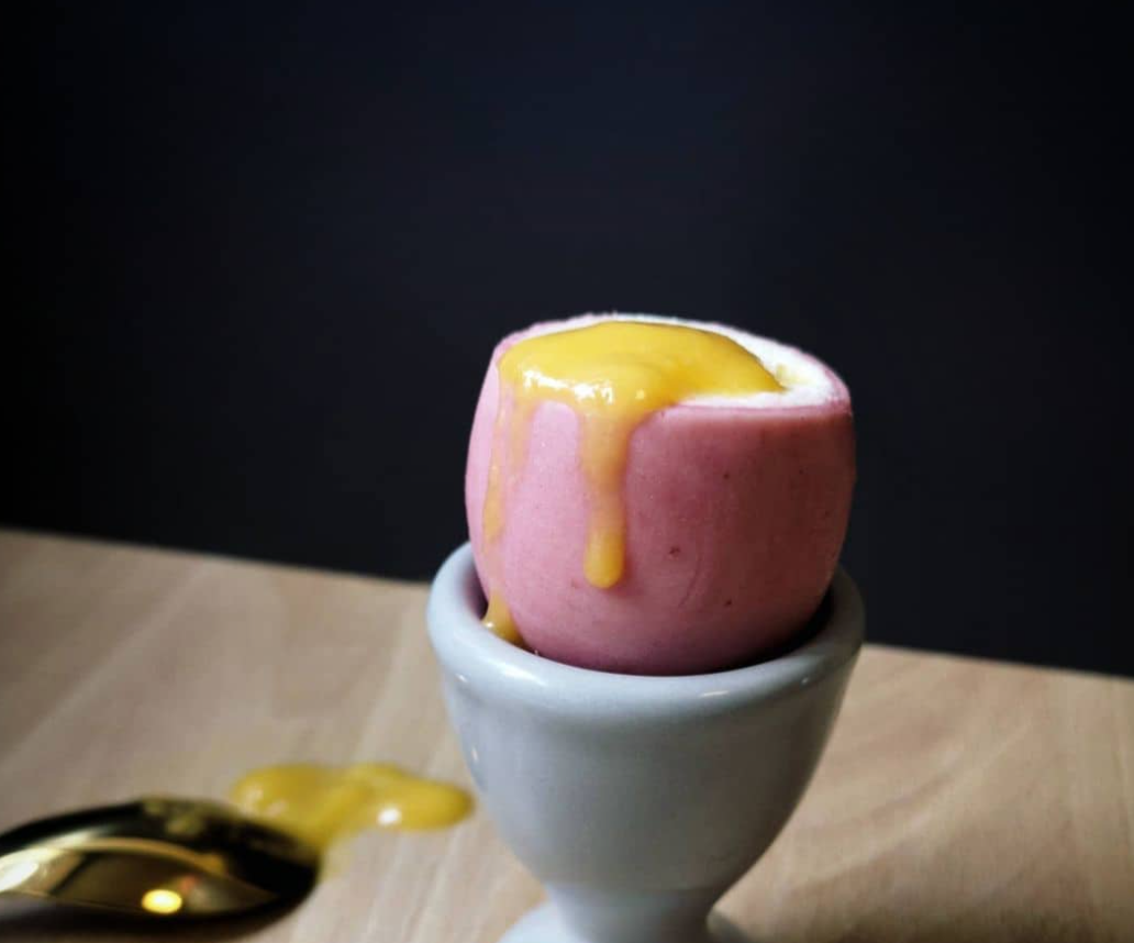 Pink softboiled egg in an egg cup with yolk spilling out on a table, dessert illusion by chefbenchurchill on instagram