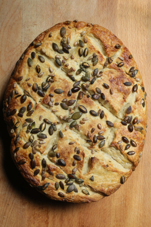 Stock photo of homemade loaf of bread with sunflower, rye and pumpkin seeds on top. This white bread is freshly baked dough in slow cooker crock pot instead of oven, organic granary crusty loaf on wooden bakery board with diagonal lattice pattern on crust