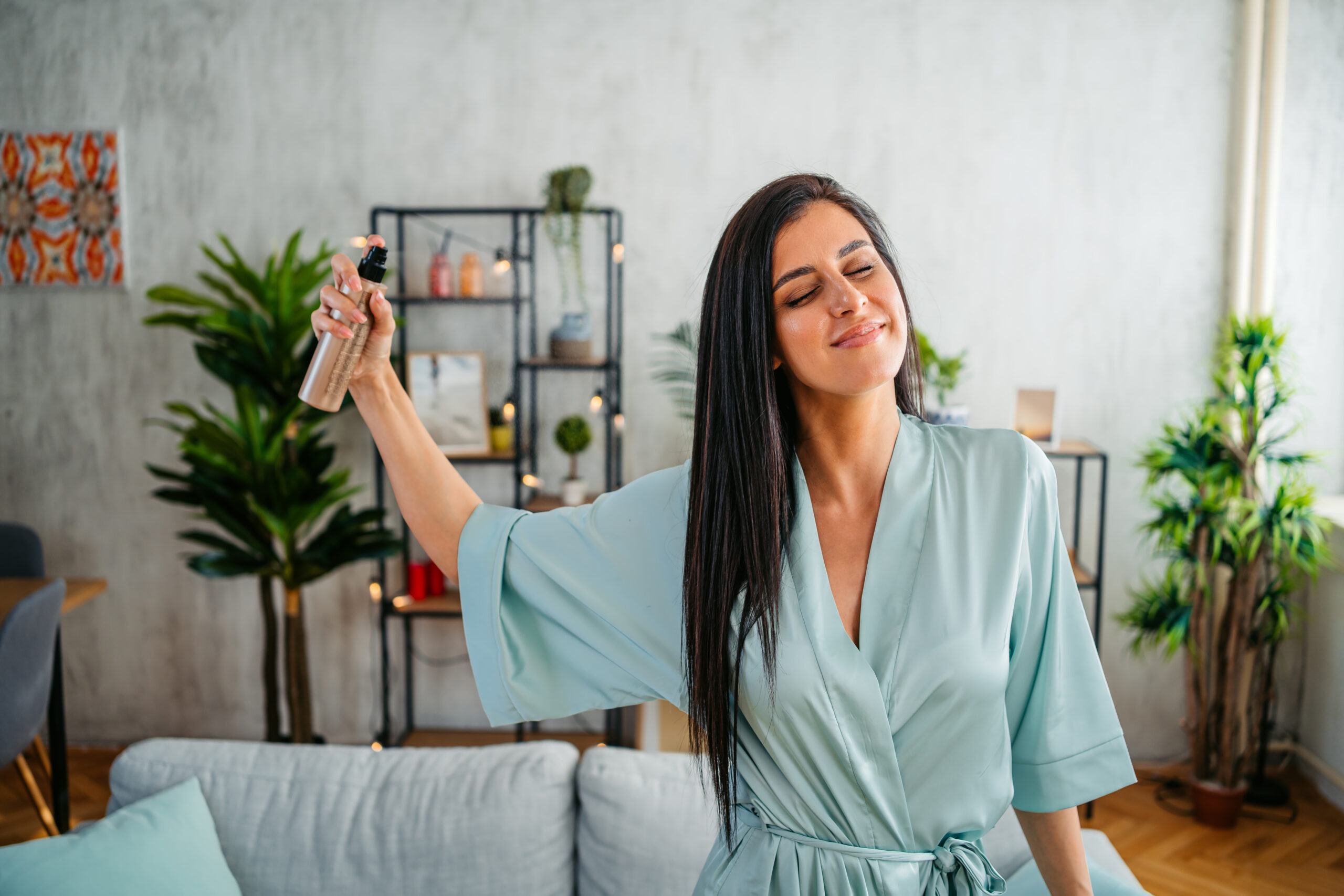 Beautiful young woman in a bathrobe using a spray bottle to spay her hair in the living room.