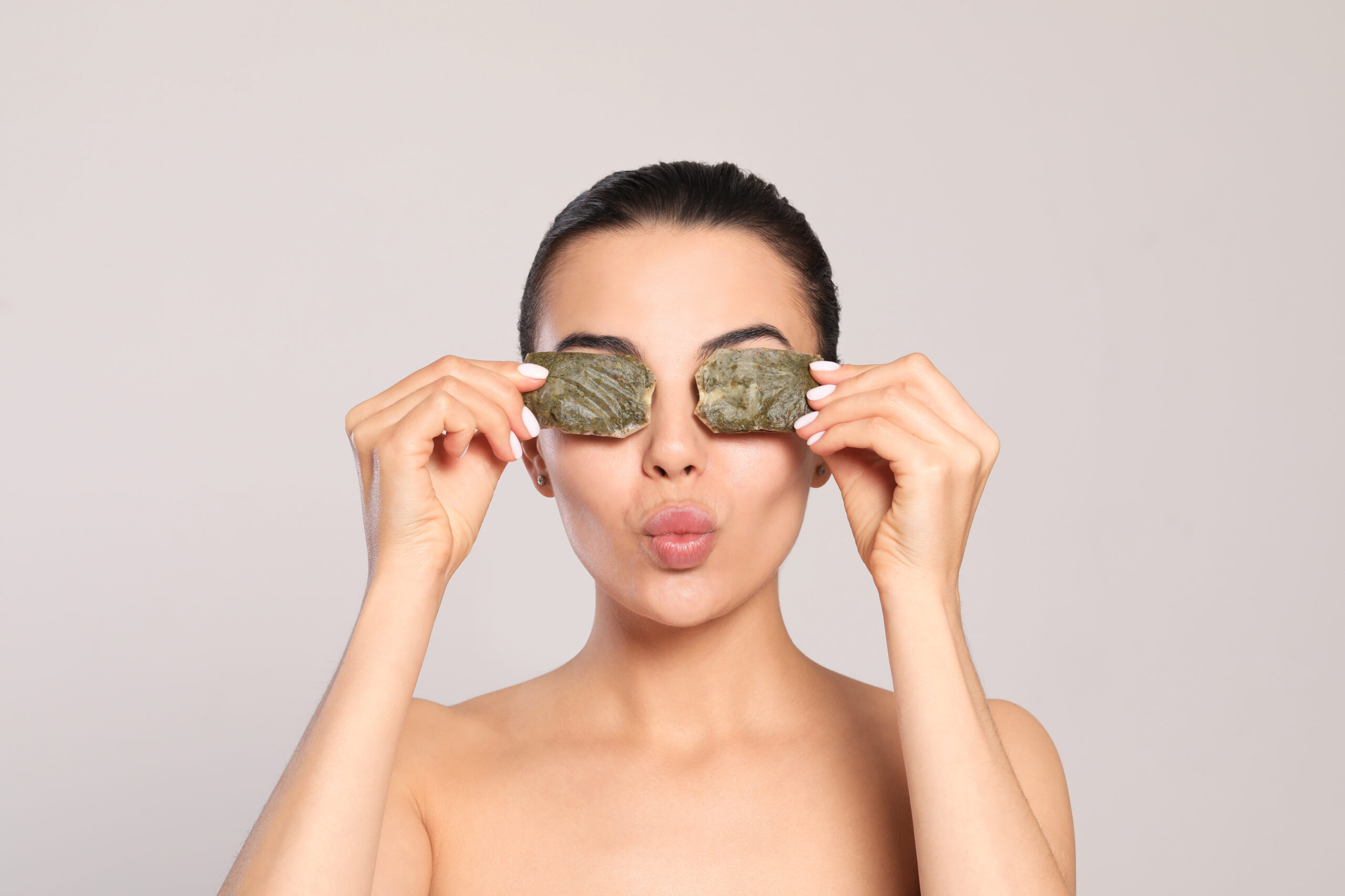 Beautiful young woman putting green tea bags on eyes against light grey background