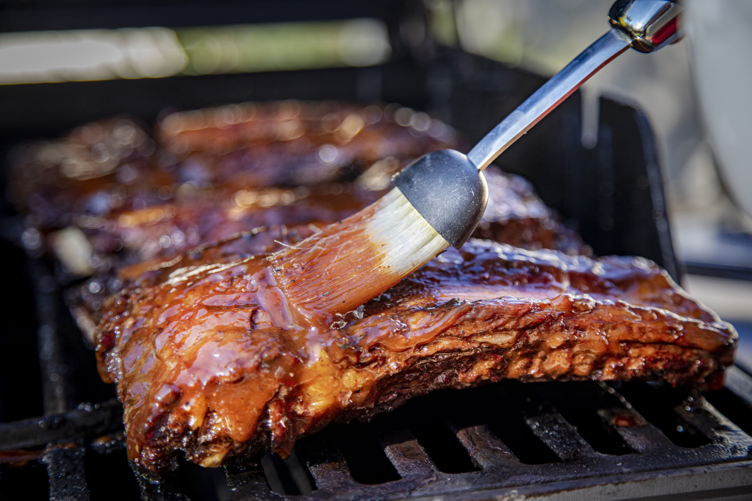 Beef ribs being basted with bbq sauce on a grill with a basting brush