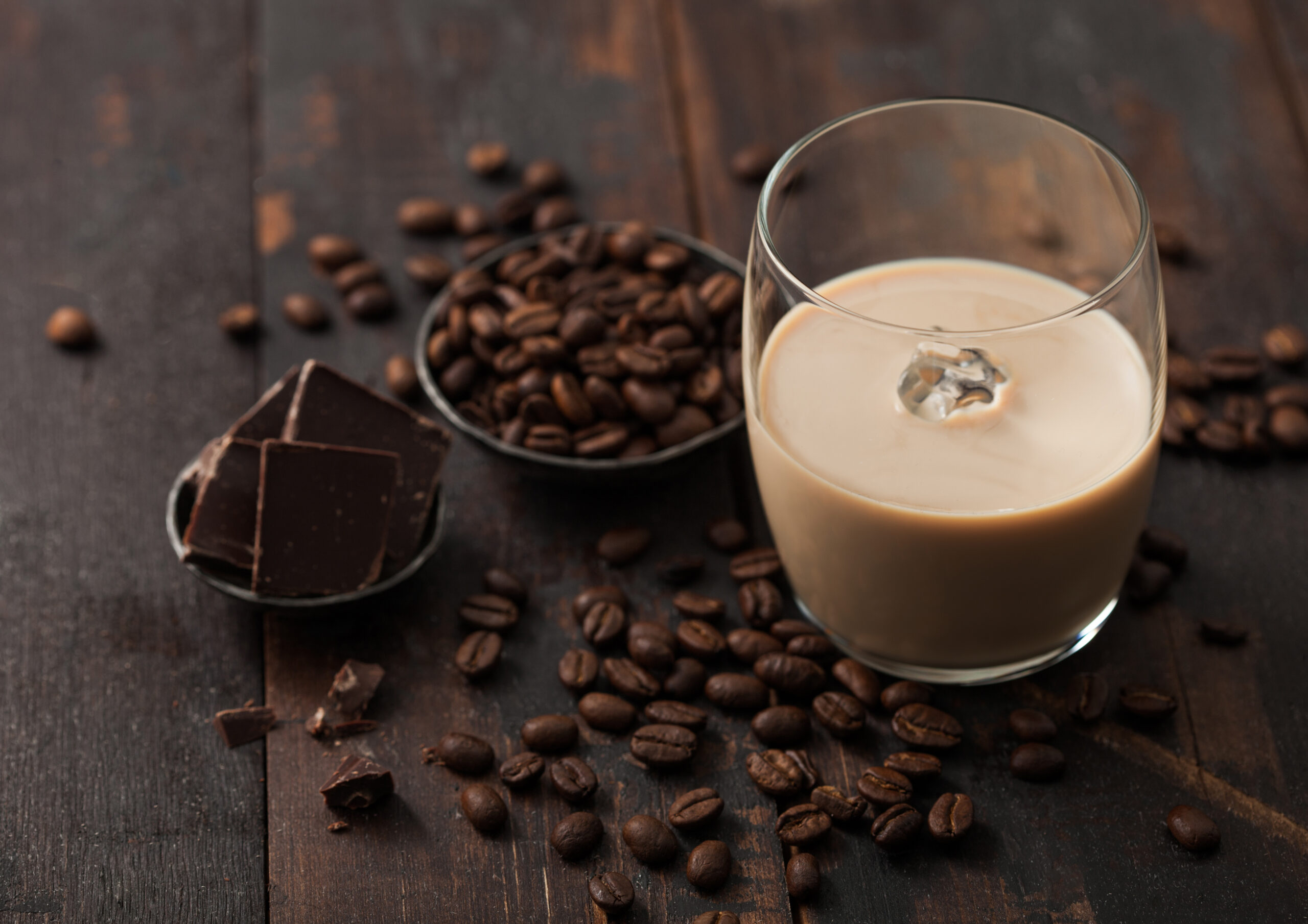 Glass of Irish cream liqueur with coffee beans and dark chocolate in steel bowl on dark wood background. Top view