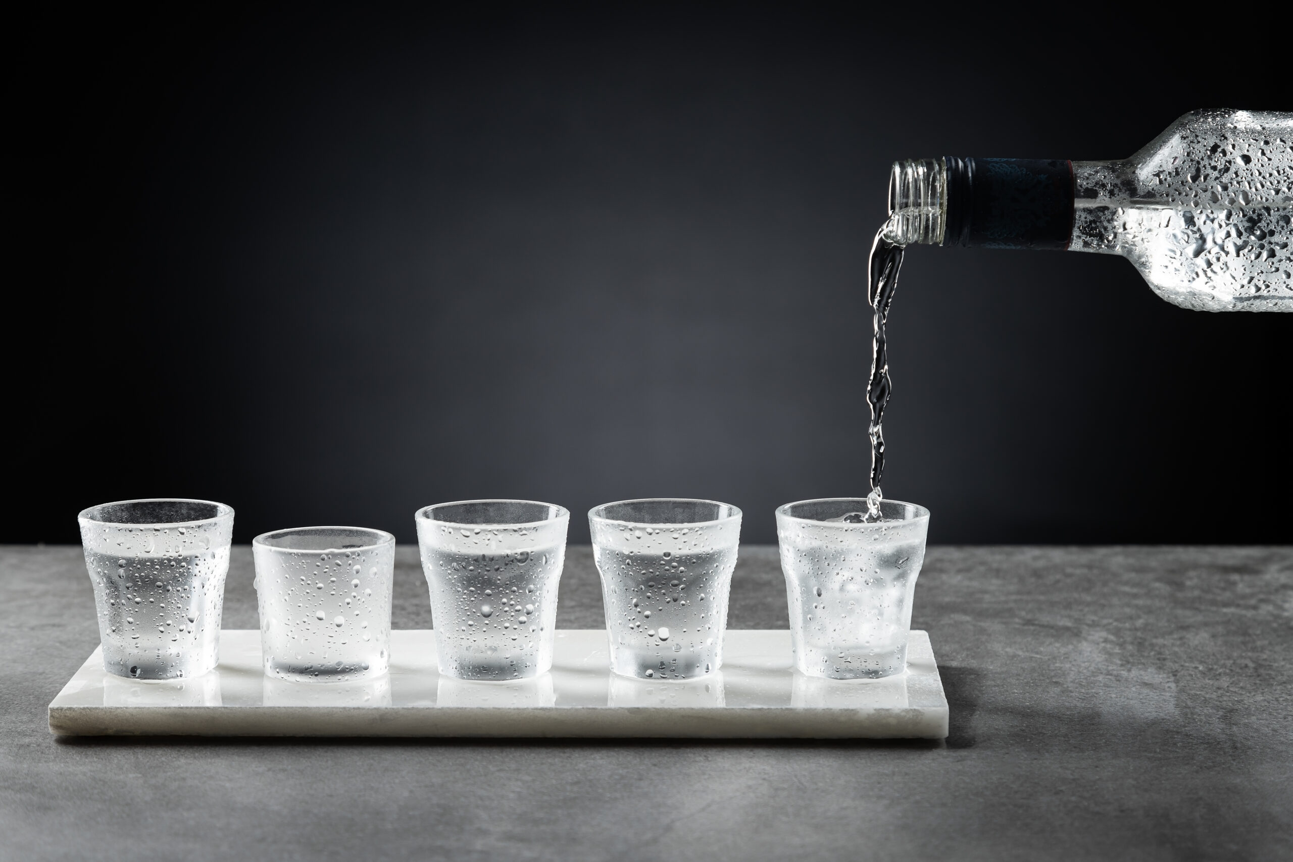 Pouring cold vodka into shot glass isolated on dark background. Selective focus.