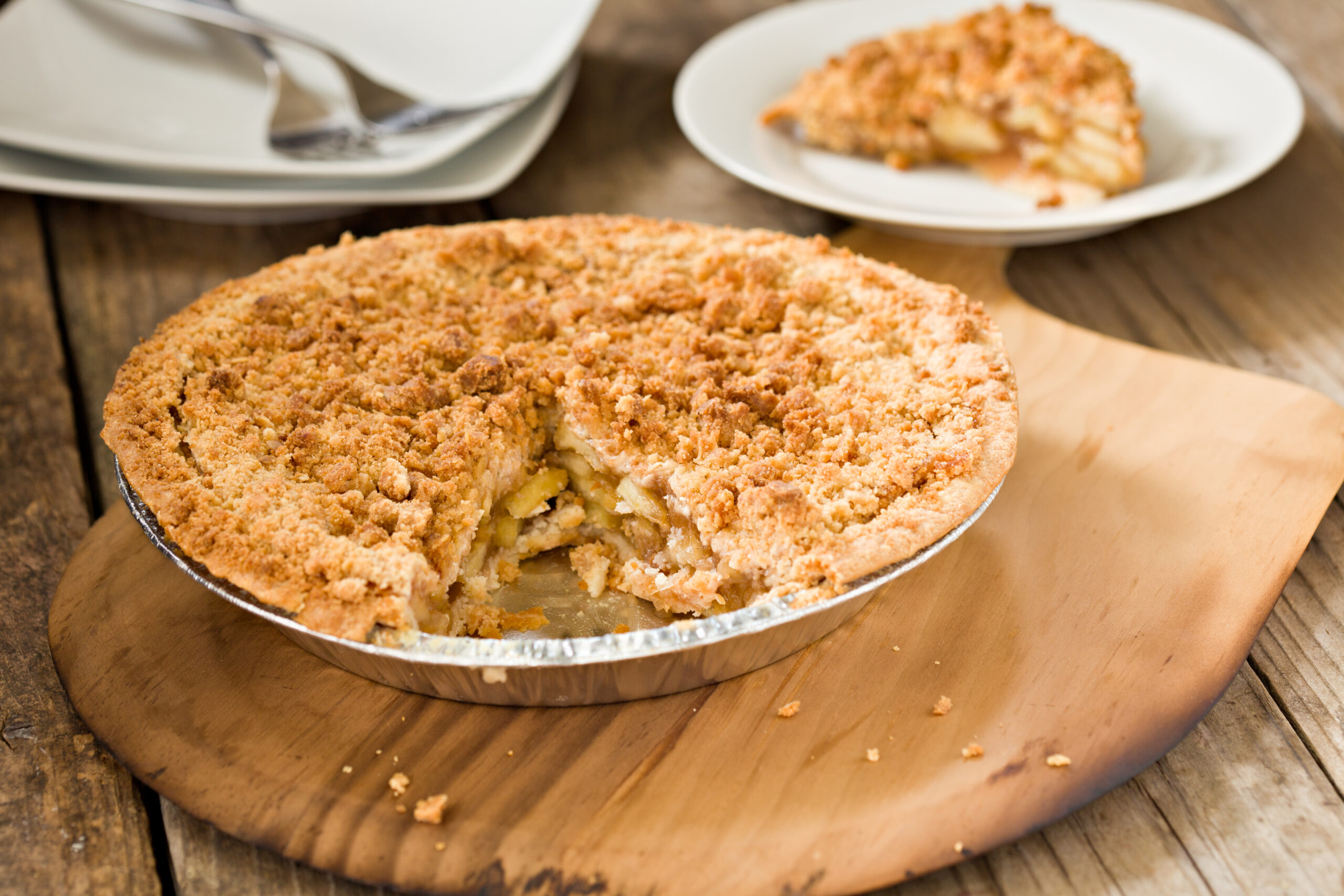 A high angle close up shot of a freshly baked dutch apple pie sitting on a wooden pizza peel and a serving of pie in the background. Shot on a grungy  old wooden table.