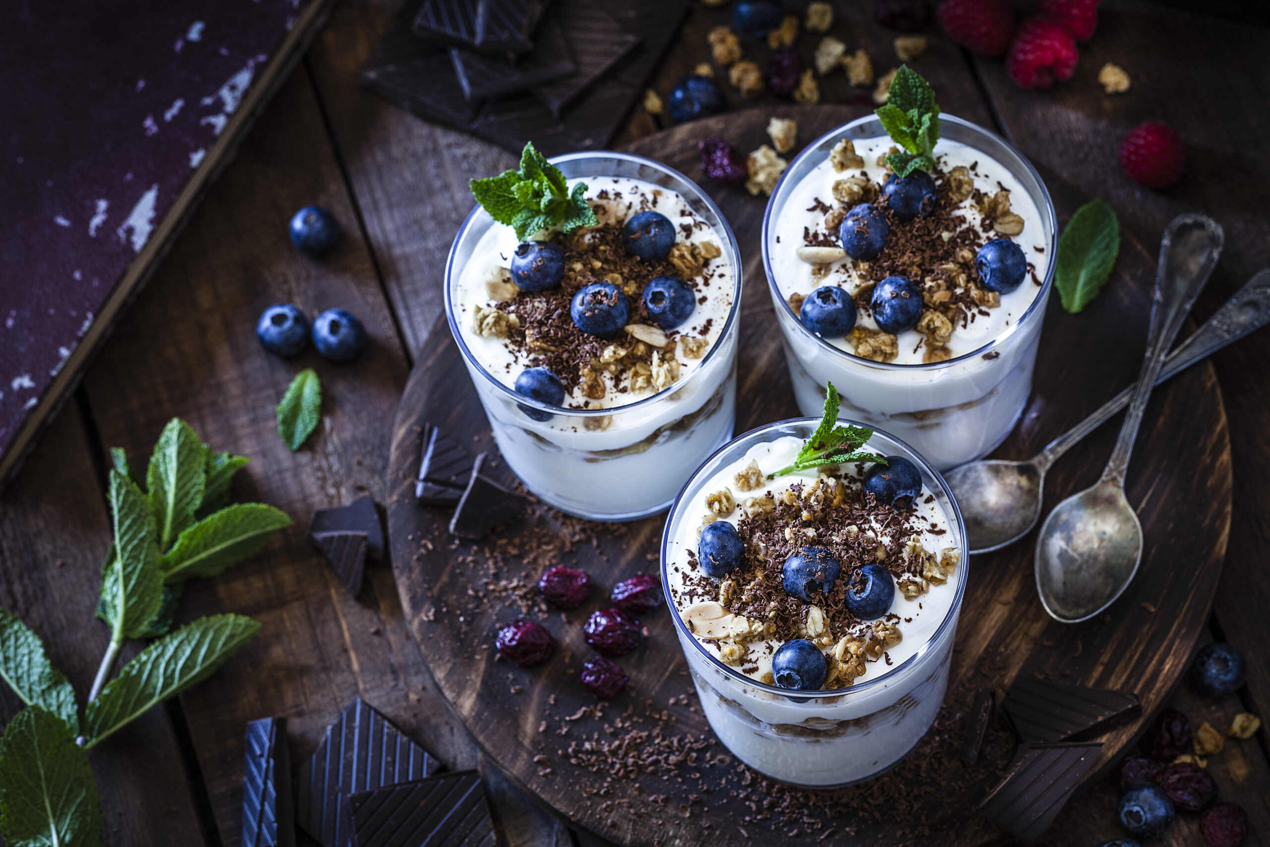 High angle view of three glasses of homemade yogurt with granola, berry fruits and chocolate shot on rustic wooden table. The glasses are on a round wooden tray and two spoons are also on the tray. Low key DSRL studio photo taken with Canon EOS 5D Mk II and Canon EF 100mm f/2.8L Macro IS USM