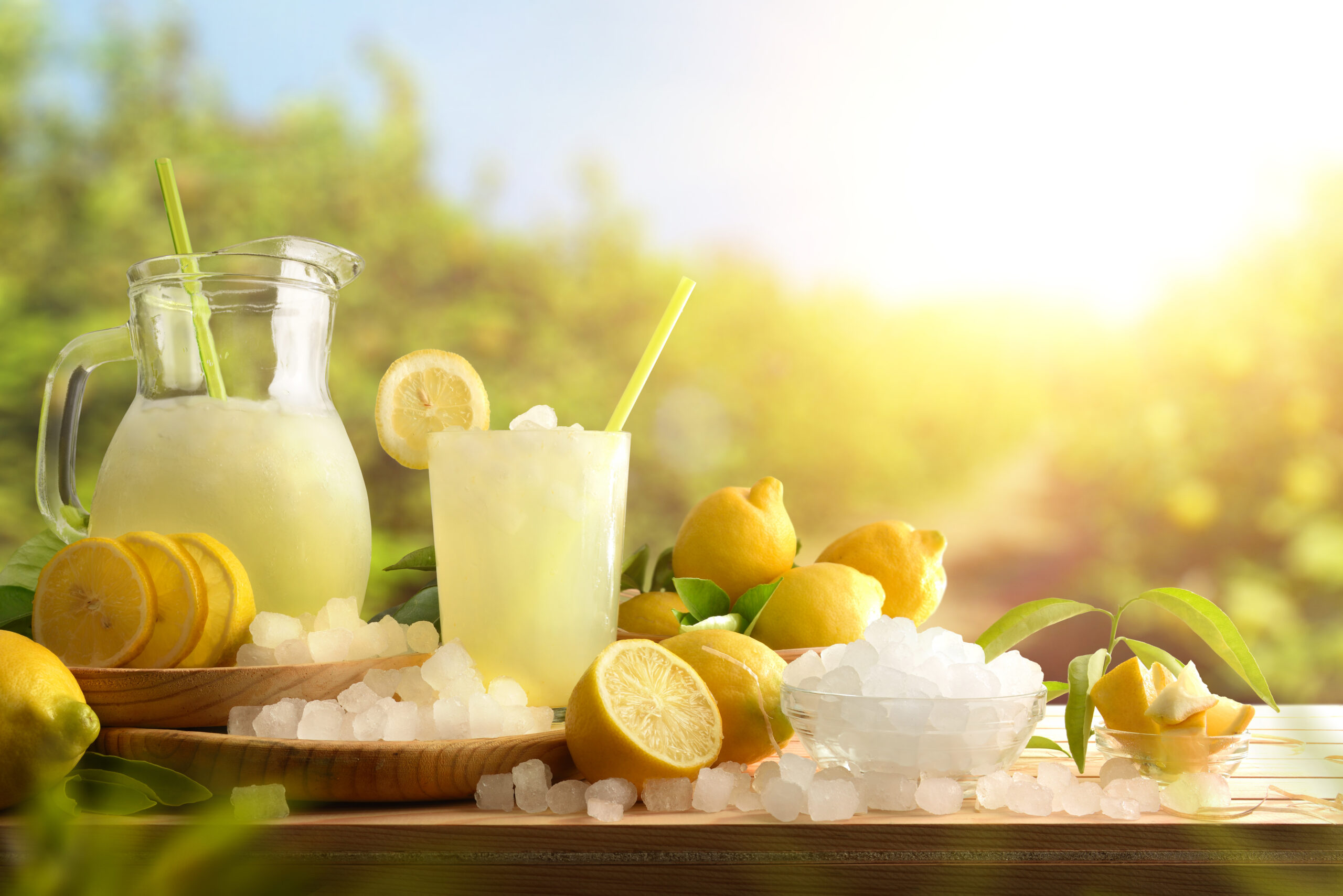 Lemonade with ice in a pitcher and glass on a wooden table with fruit and crushed ice outside with a lemon field in the background on a sunny day. 