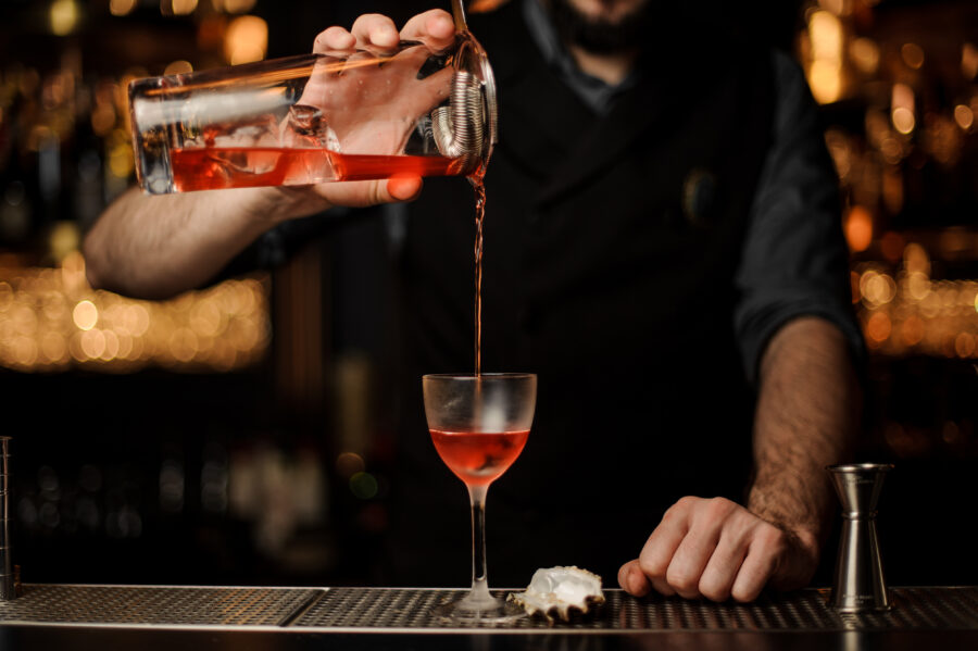 Male bartender in gray shirt pours fresh and tasty alcohol cocktail using jar with steel sieve