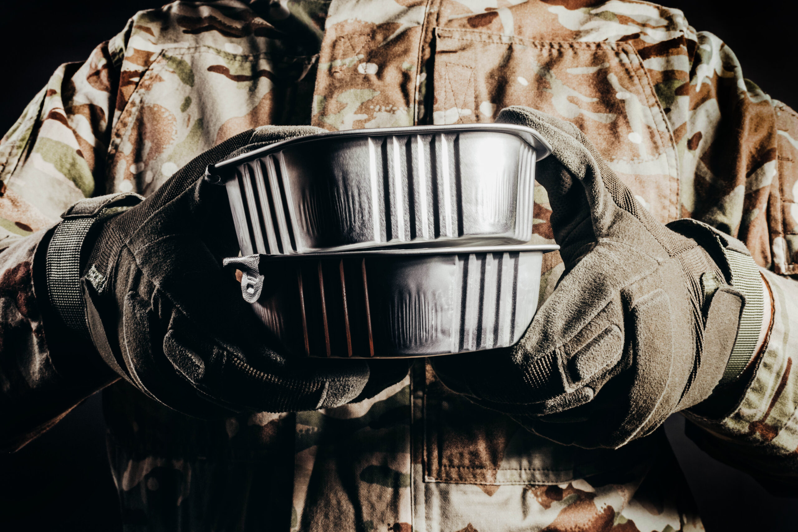 Photo of soldier in camouflaged uniform and tactical gloves holding canned food MRE on black background, close-up view.