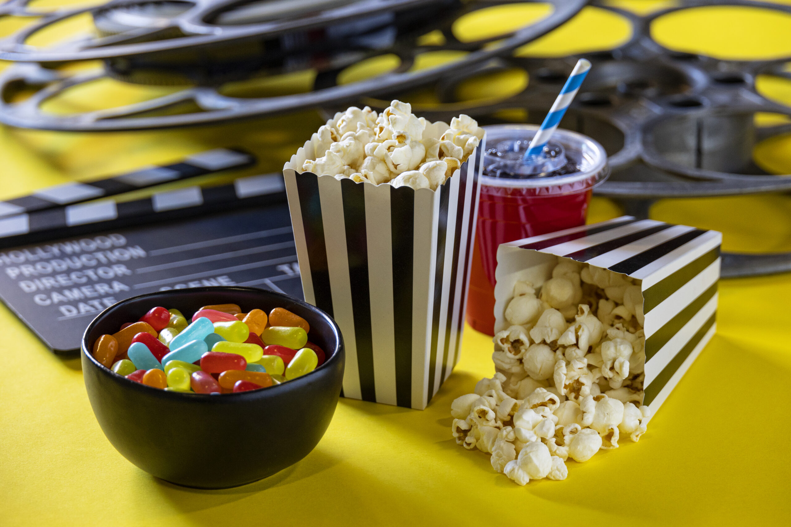 This is a conceptual photo relating to going out to the movies or watching a movie at home. There are two old retro movie reels on a bright yellow background with two bags of popcorn and hand made cinema tickets.