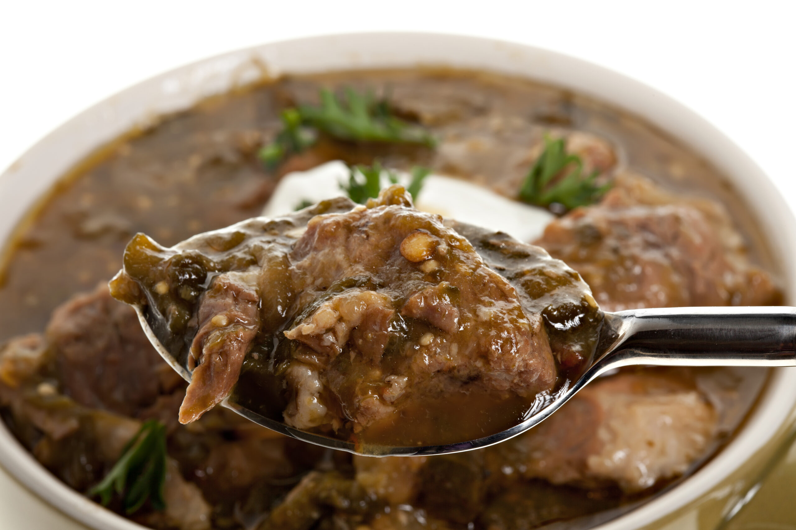 A macro of chili verde in a spoon with a bowl of it in the background on a pure white background.