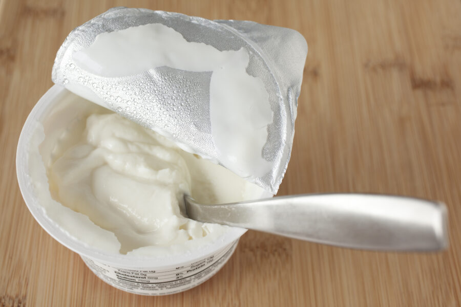 A small container of greek yogurt with the spoon in it and the foil covering pulled back.