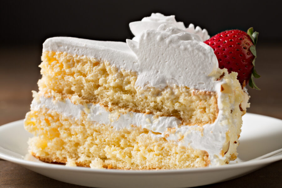 An extreme close up horizontal photograph of a side view of a slice of tres leche cake topped with white cream and decorated wit a ripe red strawberry.