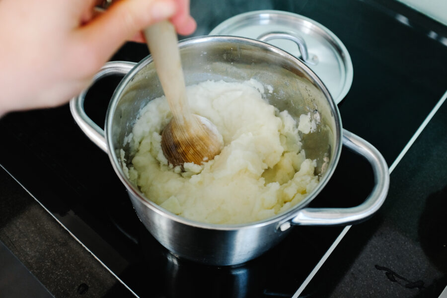 preparing mashed potatoes in real kitchen - hand mashing potatoes with wooden masher inside metal cooking pan, close up view, real life, cooking stove