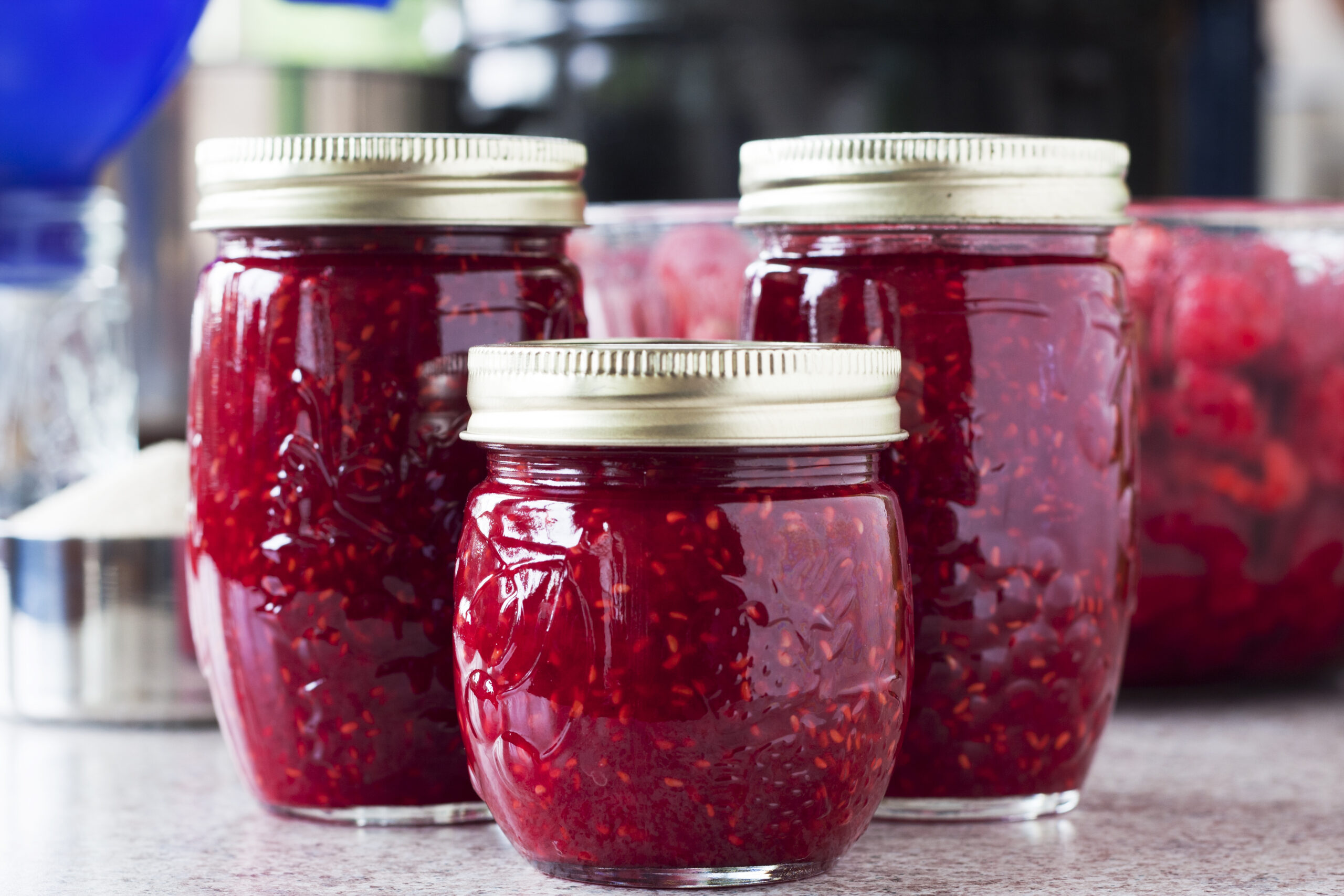 Three completed jars of jam, a bowl full of fresh rasperries, a measuring cup full of raw sugar, an empty jelly jar with a canning funnel, and pans in the background