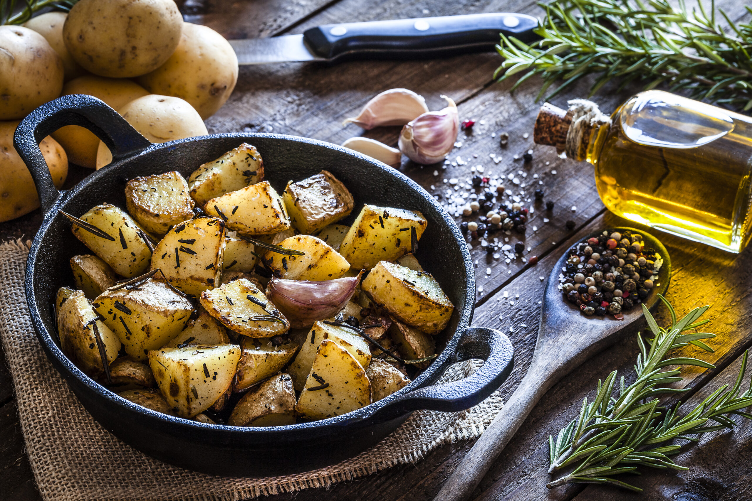 Cast iron pan filled with roasted potatoes shot on rustic wooden table. The cooking pan is at the left of an horizontal frame and the ingredients for cooking the potatoes are all around the pan placed directly on the table. The ingredients includes are raw potatoes, rosemary, olive oil, salt, pepper and garlic. Predominant colors are brown and yellow. DSRL studio photo taken with Canon EOS 5D Mk II and Canon EF 100mm f/2.8L Macro IS USM