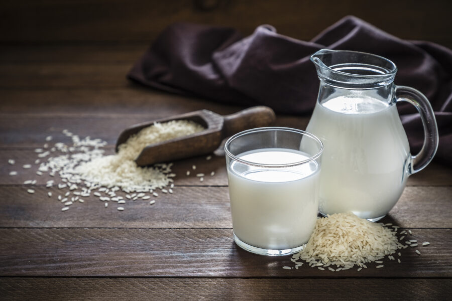 Front view of a jug and a drinking glass full of rice milk. The glass and the jug are at the right side of the image on a rustic wooden table, next to the glass its a heap of rice grains. 