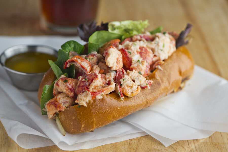 Lobster roll. Maine lobster mixed with mayo, celery, onions, garlic, scallions, chives, lemon juice. Lobster roll on toasted hotdog bun w/ lettuce, tomato, garlic mayo seasoned salt & pepper. Classic American restaurant or diner lunch sandwich favorite.