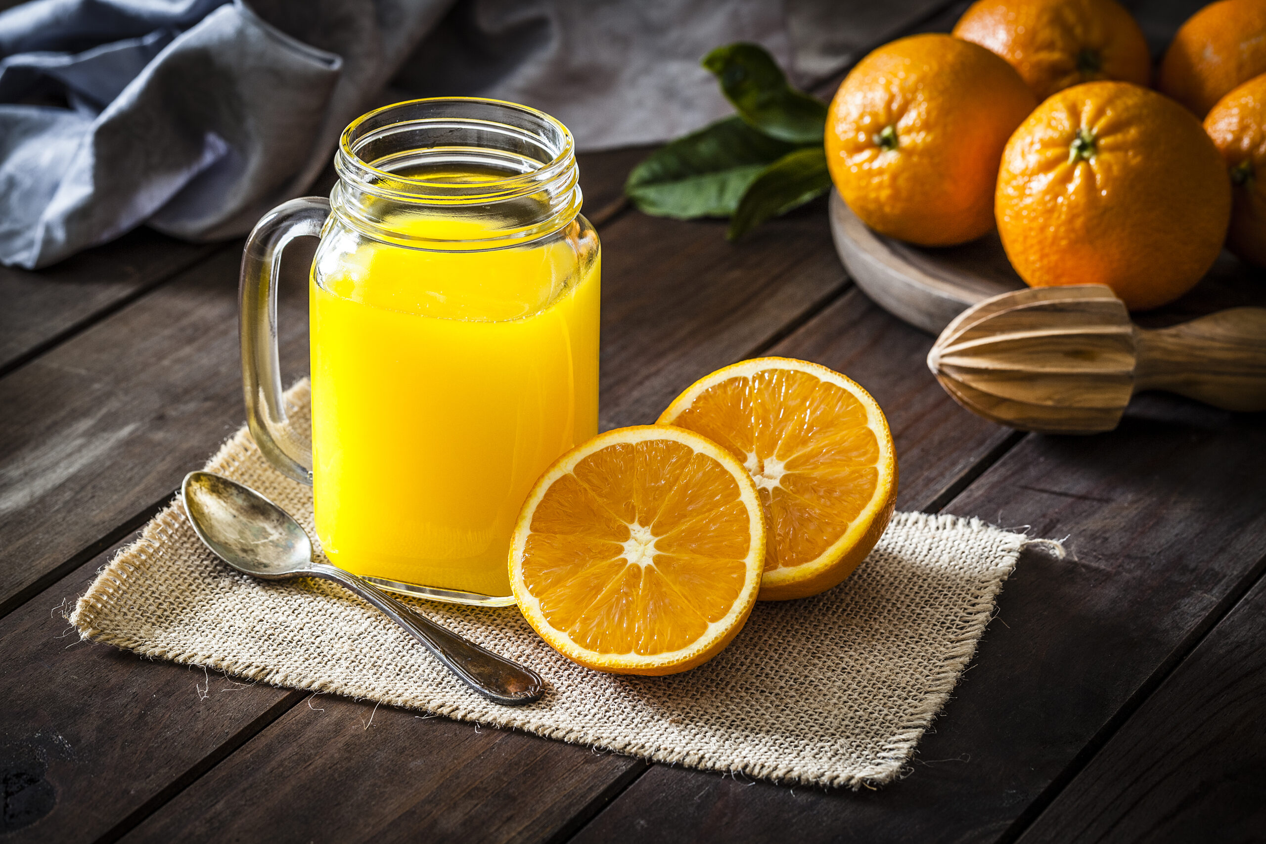 Orange juice glass jar shot on rustic wooden table. The jar is on a burlap cloth and two orange halves are beside it. An old metal spoon and a wooden juicer complete the composition. A round wooden tray with fresh oranges is at the top-right corner of an horizontal frame. Predominant colors are orange and brown. DSRL studio photo taken with Canon EOS 5D Mk II and Canon EF 100mm f/2.8L Macro IS USM