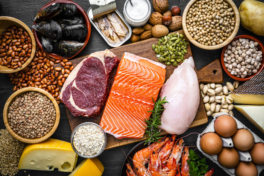 Overhead view of a large group of food with high content of healthy proteins. The composition includes salmon beef fillet, chicken breast, eggs, yogurt, mussels, chick peas, pistachios, cheese, brown lentild, beans, shrimps, canned tuna, pumpkin seeds, soybeans among others. 