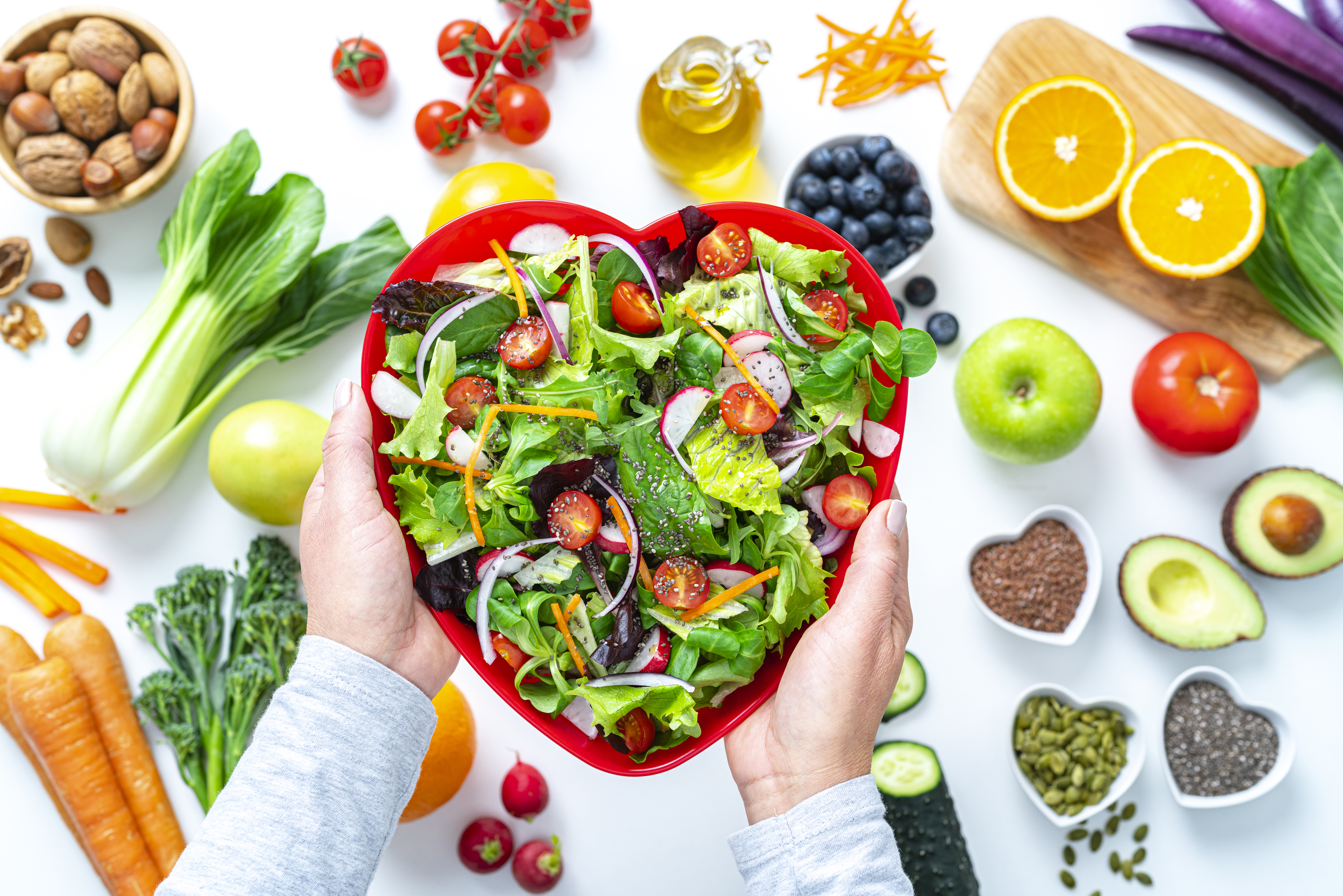 Overhead view of female hands holding a red heart shape plate with healthy salad shot on white background. Multi colored fresh fruits, vegetables, seeds and nuts are out of focus at background