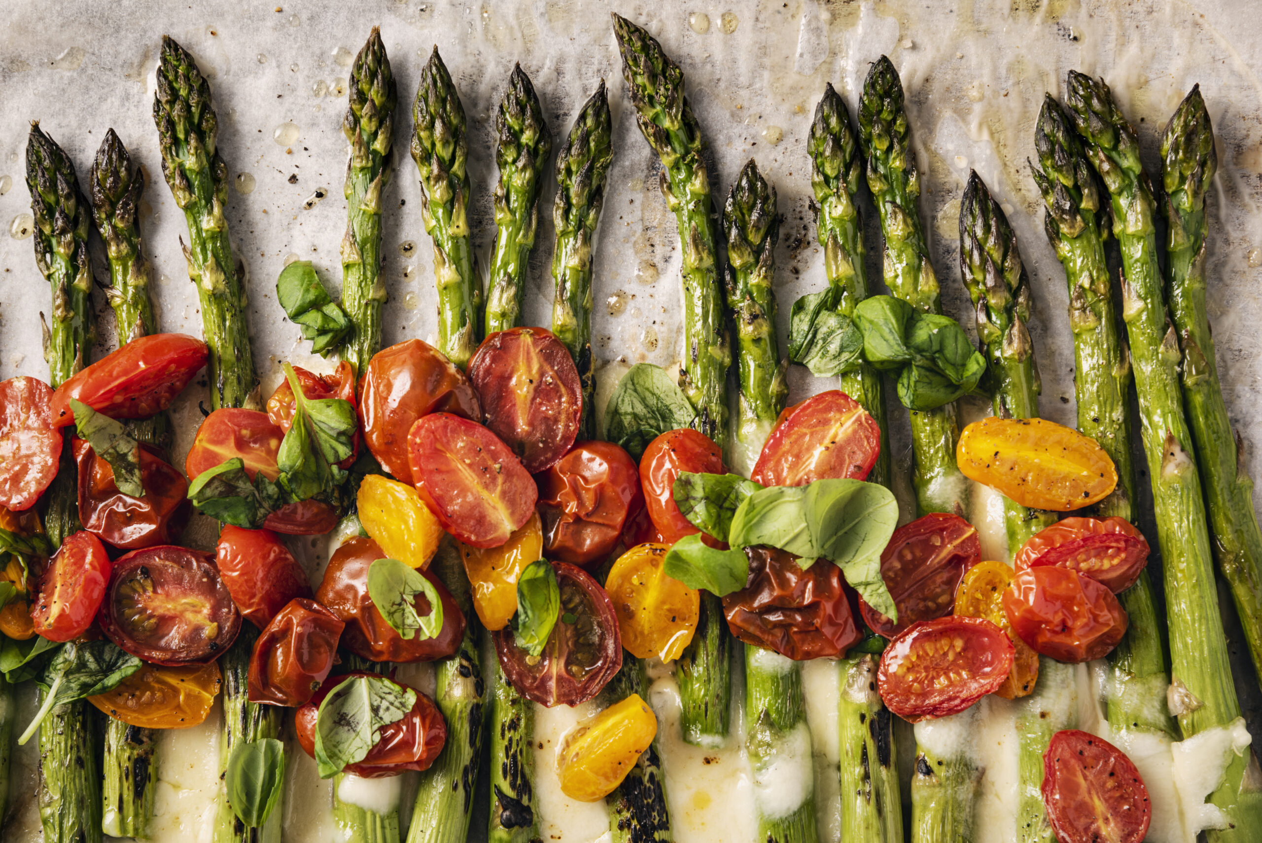 Remove the last few woody centimetres of the asparagus stalk, place on lined baking tray with the halved tomatoes, drizzle with olive oil and season with salt and pepper and bake at 180C/375F for 8-12 minutes. For the last couple of minutes cooking time add the shredded mozzarella to melt over the asparagus. Before serving, mix the tomatoes with the asparagus and melted cheese and add some fresh basil leaves. Colour, horizontal with some copy space.