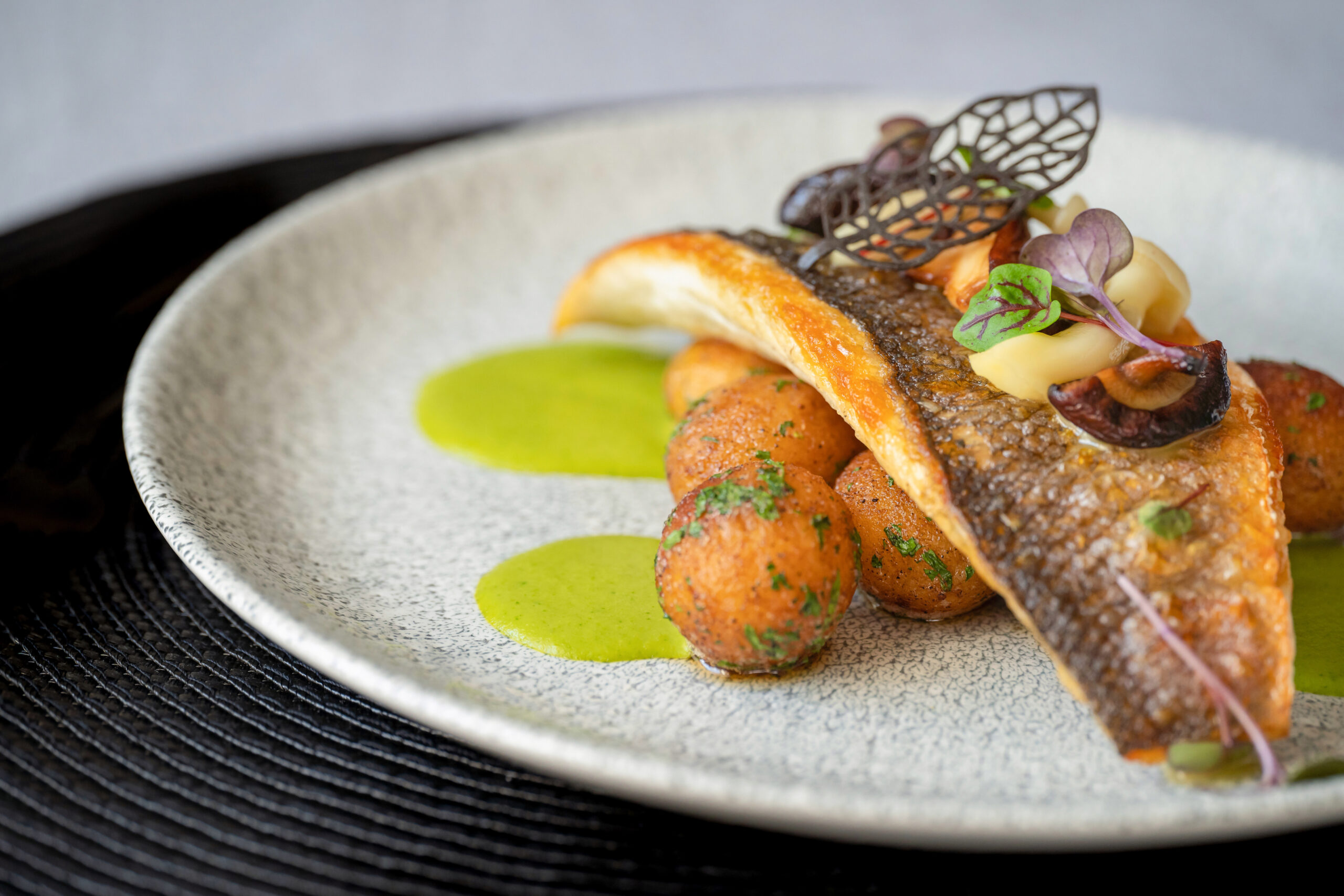 Roasted branzino fish filet with marinated mushrooms, asparagus sauce and butter potatoes on the plate on black tablecloth. Gourmet food. Chocolate leaf decoration.