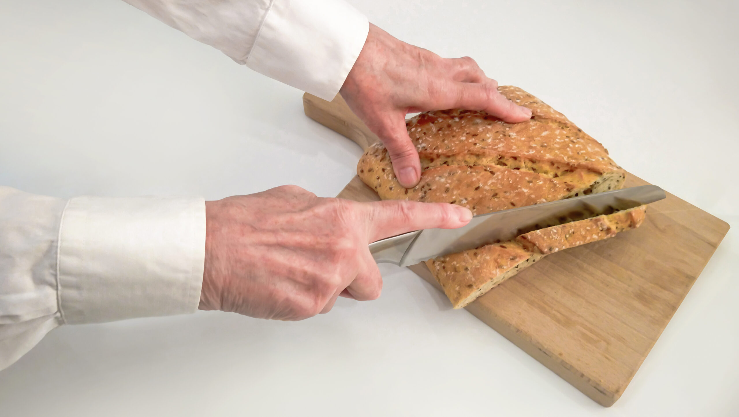 Senior female hands cutting integral whole grain bread loaf with large stainless steel bread knife, on wooden chopping board, set on neutral white background, high resolution stock photo.