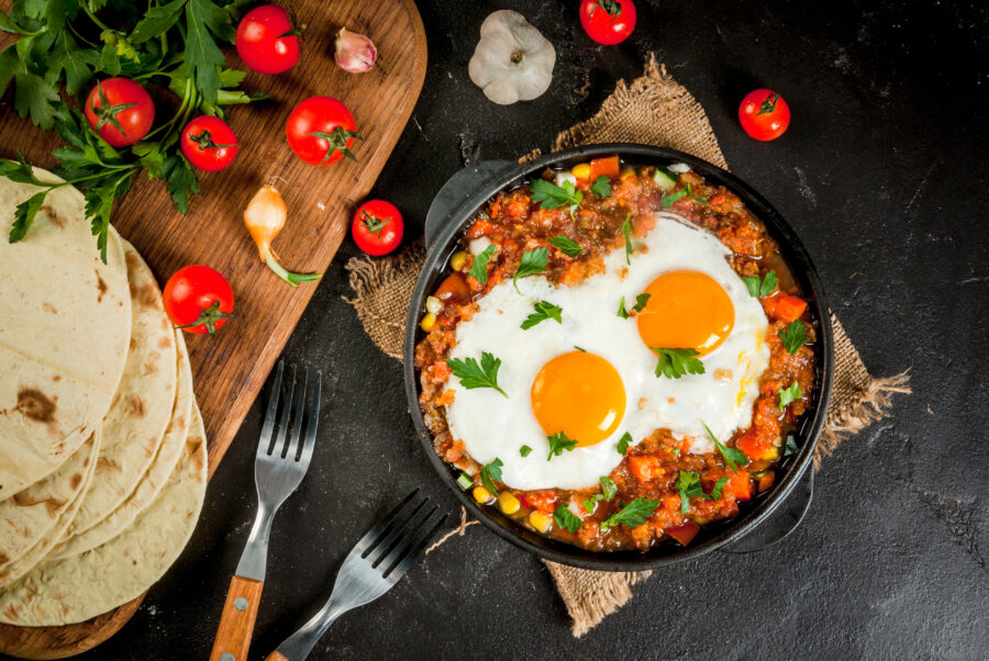 Traditional Mexican dish Huevos rancheros - scrambled eggs with tomato salsa, with taco tortillas, fresh vegetables and parsley. Breakfast for two. Top view. With a forks, copy space