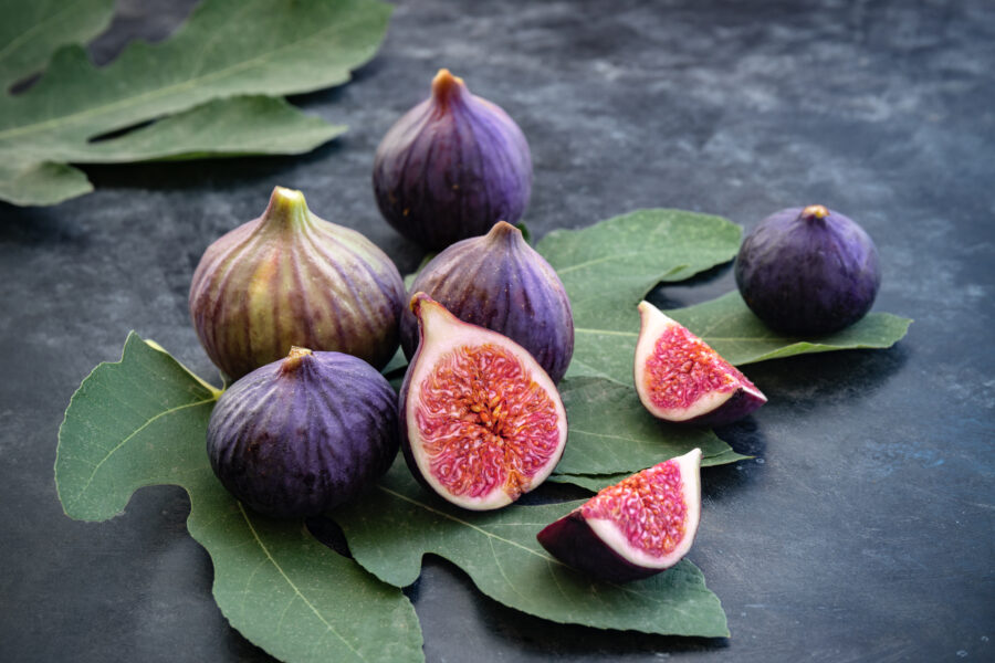 Whole and sliced figs on big fig leaves. Fresh ripe sweet fruits on dark background. Close-up. Selective focus. Blurred background.