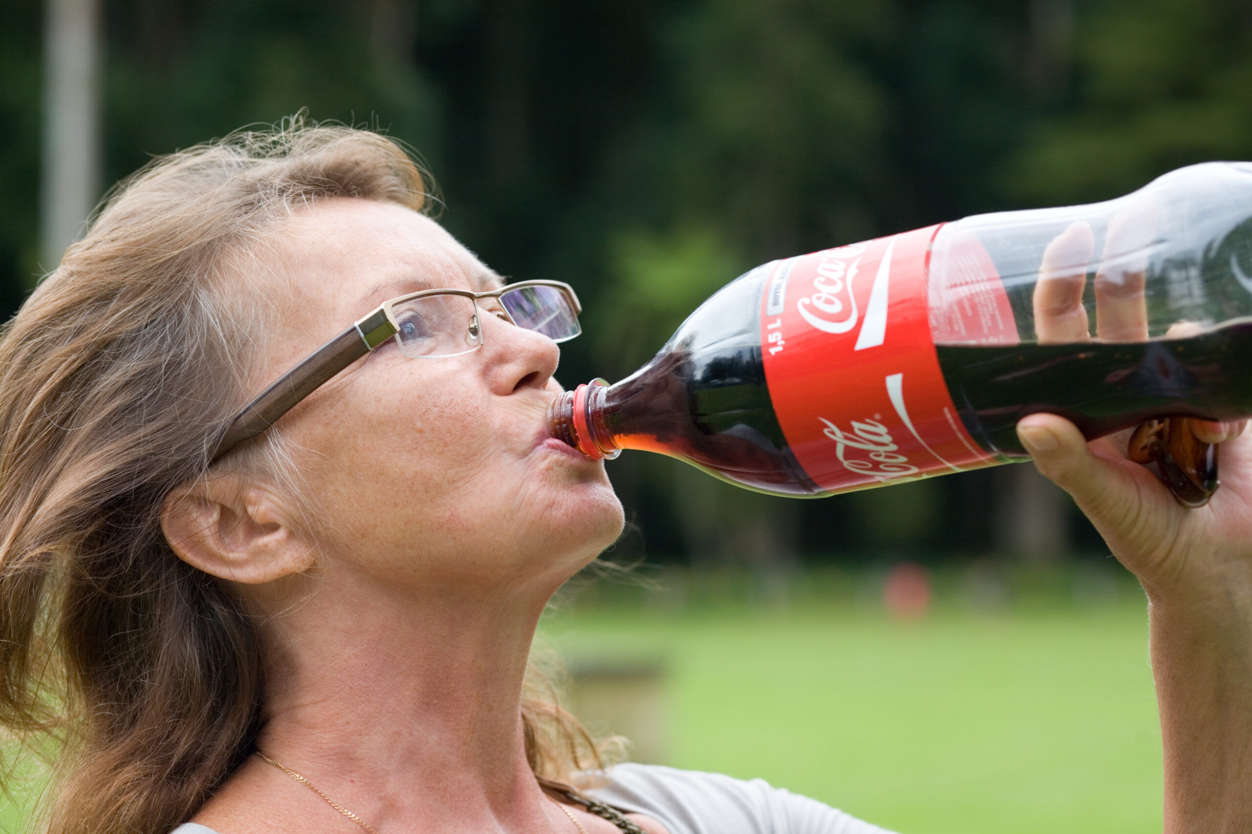 senior woman drinking Coca Cola from big bottle in park