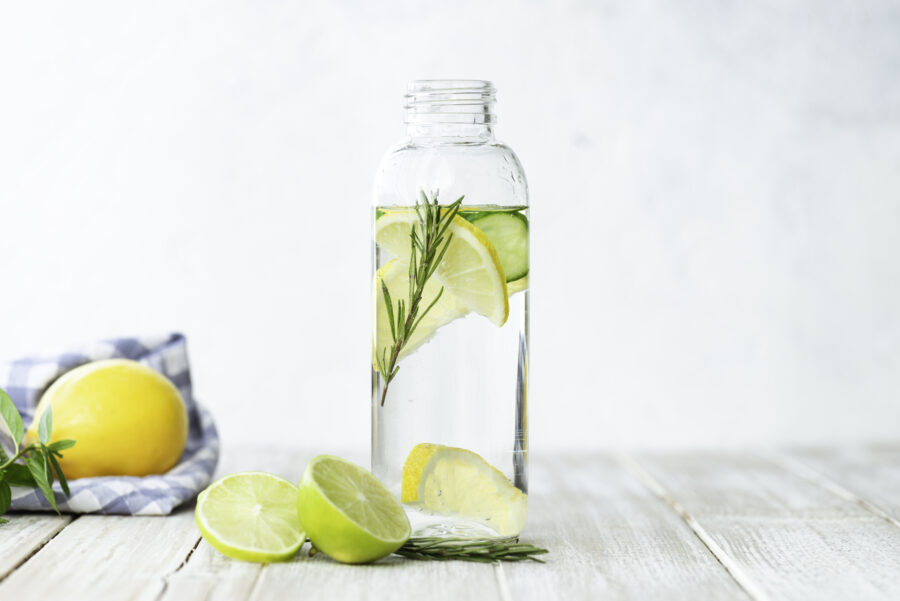 Bottle of infused water on white wood with a slice of lemon , cucumber and rosemary leaf in it.