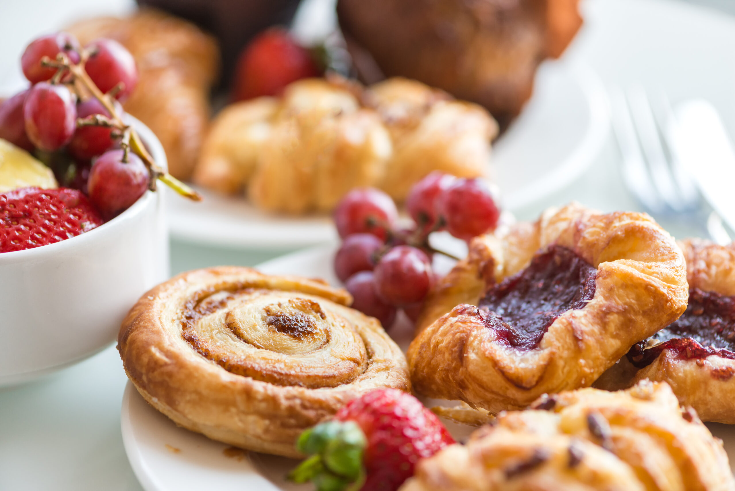 Closeup of a continental breakfast at a hotel, with bowls and plates filled with a selection of breakfast bakery items and fresh fruit on a white table with cutlery in the background.