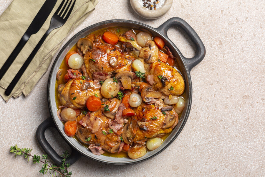 Coq au vin or rooster, cock with wine.  French dish of chicken braised with red wine, lardons, mushrooms, carrot and onion. Seasoned with salt, pepper, thyme. Beige table surface. Copy space.