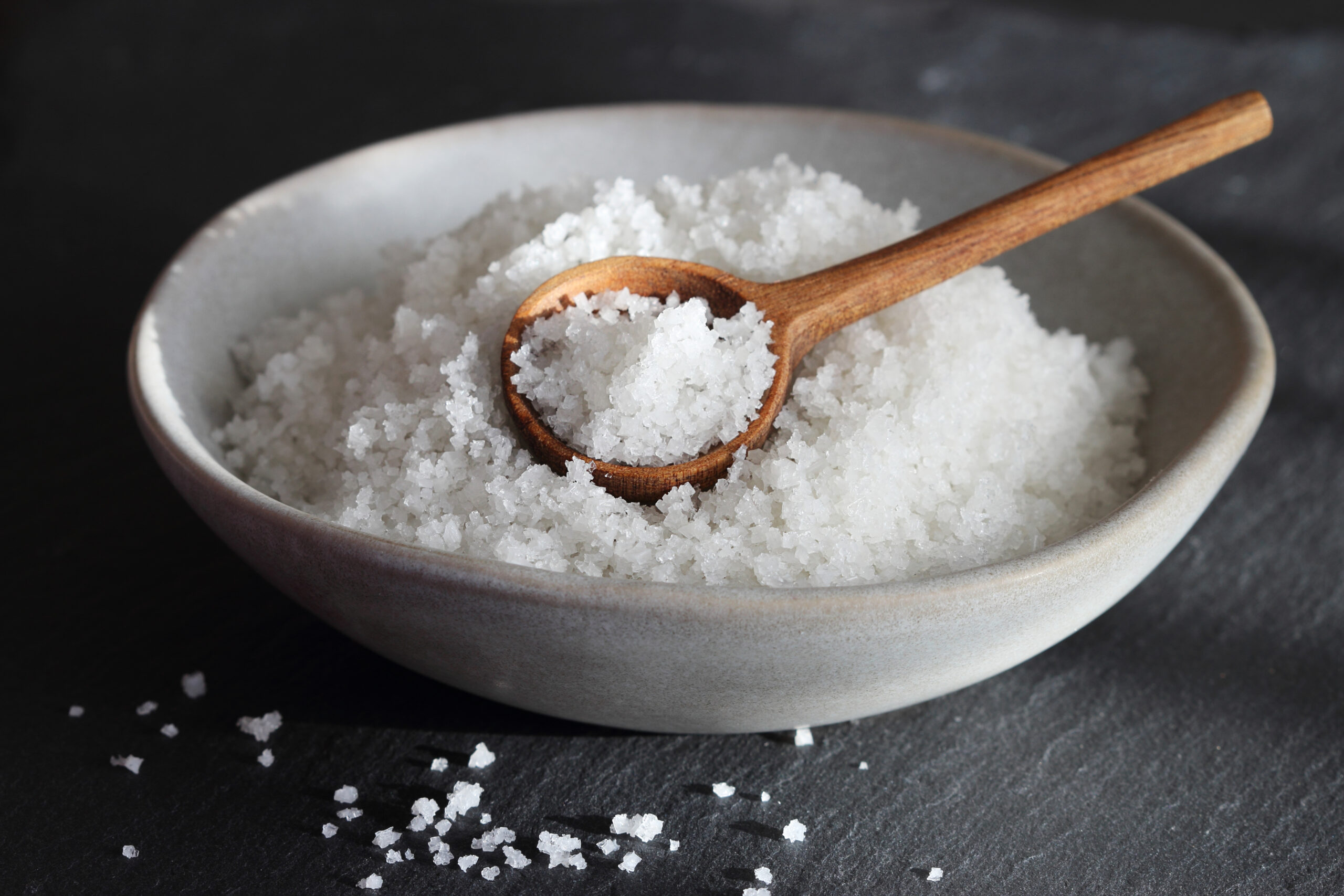 salt in a ceramic grey bowl with wooden spoon on a black slate background.