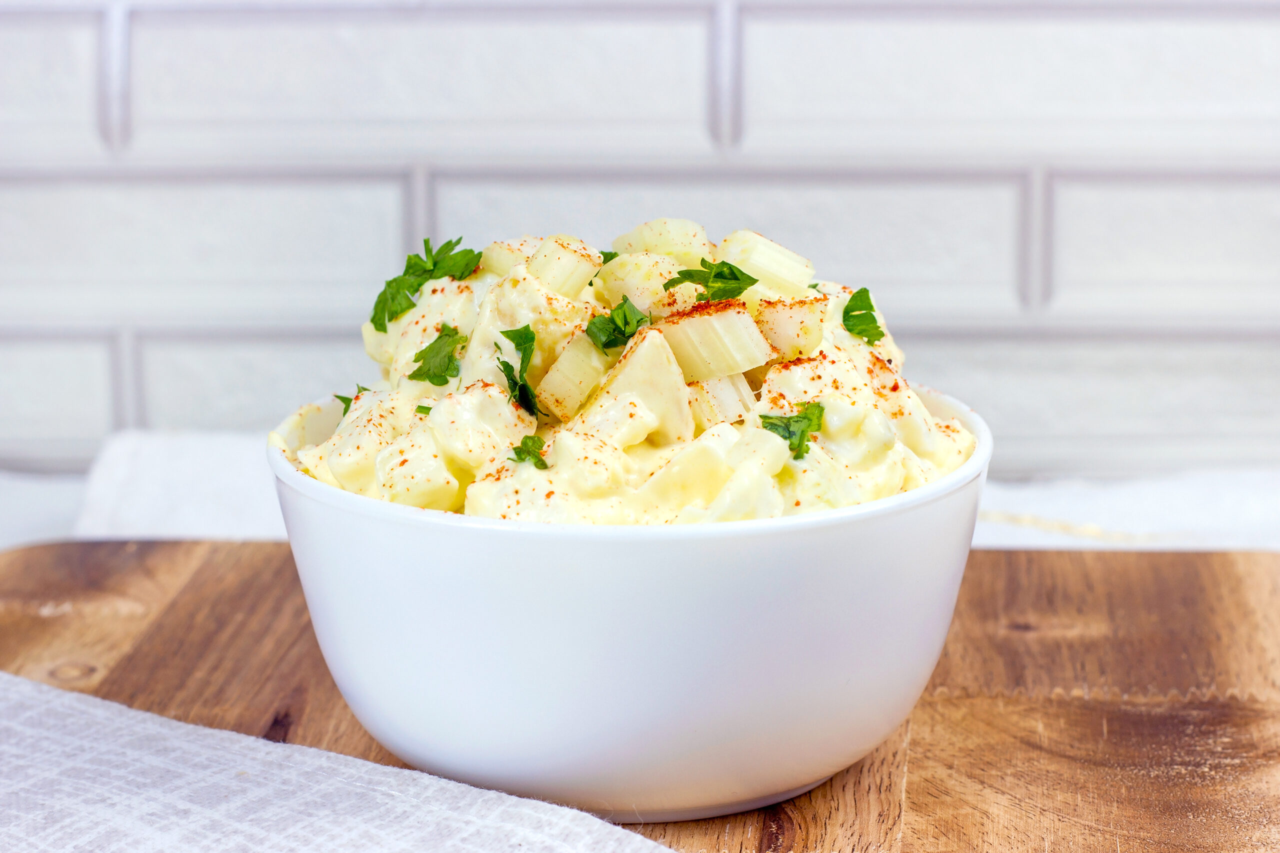 Fresh homemade traditional old-fashioned Amish Potato Salad with potatoes, boiled eggs, creamy dressing and celery in a white bowl on wooden background.